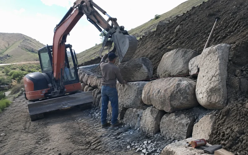 Excavator setting a large boulder into a hillside retaining wall