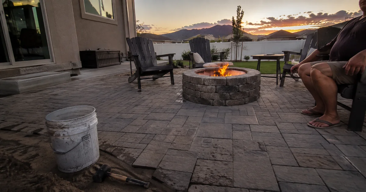 Backyard paver patio with a stone fire pit and chairs set around it at dusk