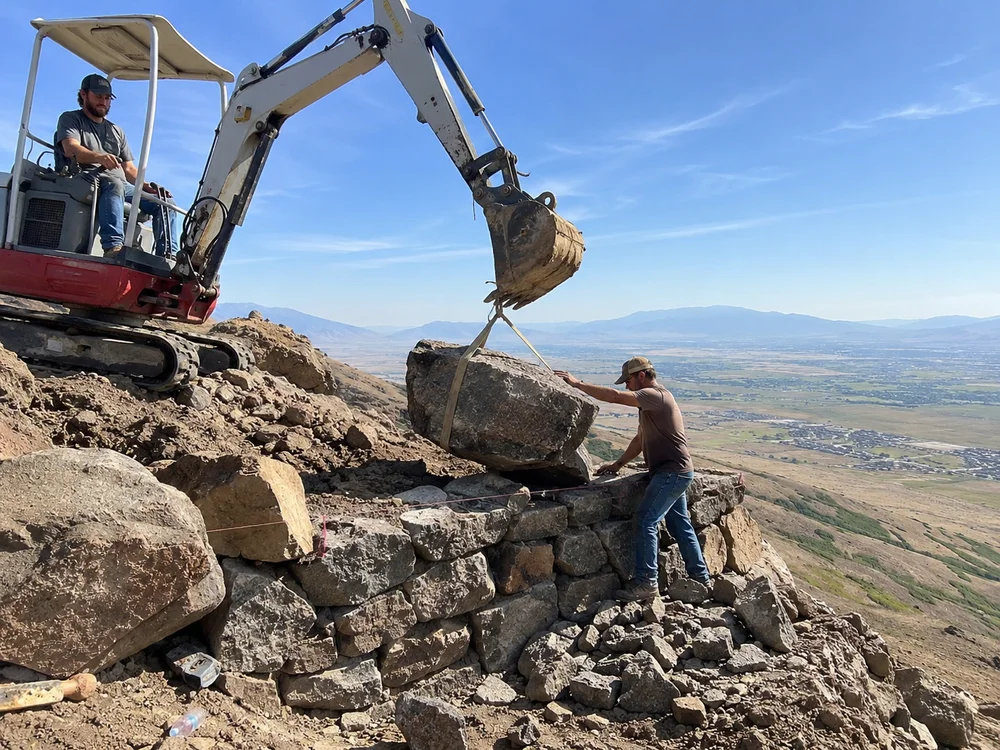 Excavator setting a large basalt boulder into a hillside wall